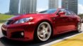 A red car driving down the street with tall buildings in the background.