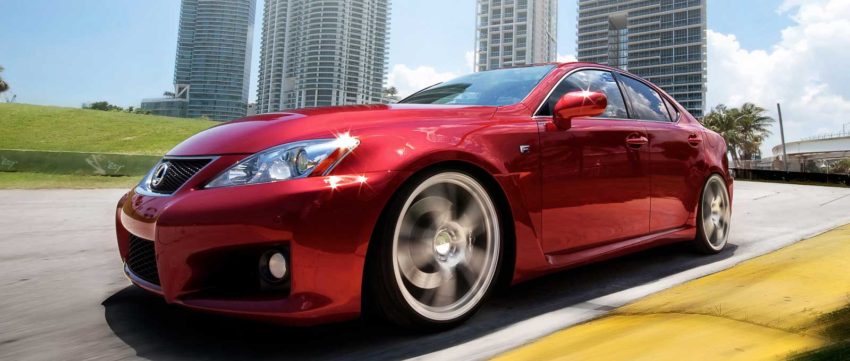 A red car driving down the street with tall buildings in the background.